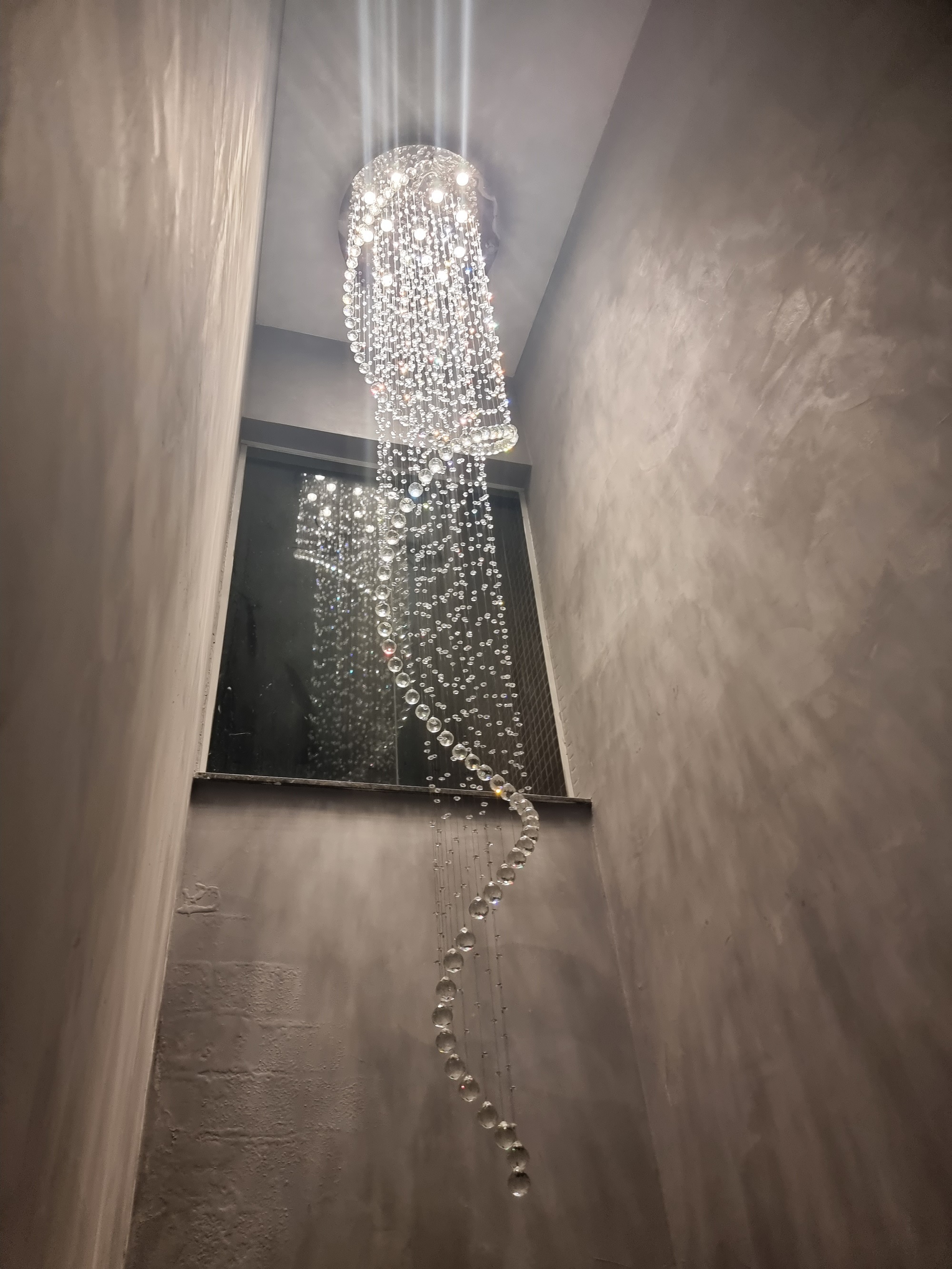 Lustre de cristal modelo "Caracol" da Linha Kria instalado em ambiente com pé direito duplo, com cristais K9 legítimos em espiral decorando sala de estar moderna.